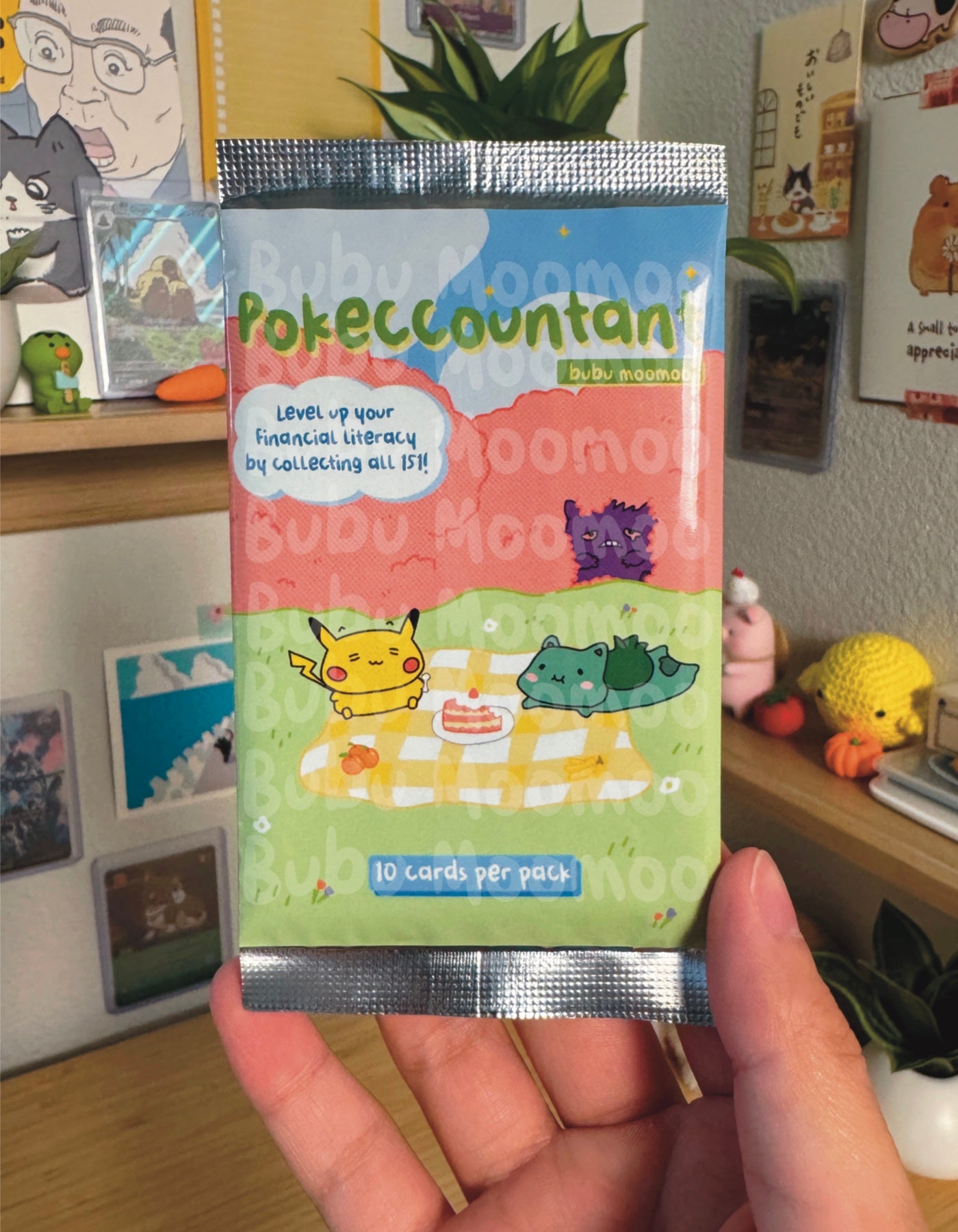 Pokeccountant collectible card pack held in front of a decorated wall with posters and toys. The Pokeccountant pack has the words "Pokeccountant" and "bubu moomoo". It also has the phrases "Level up your financial literacy by collecting all 151!" and "10 cards per pack". The pictures on the pack includes cartoon characters having a picnic on a pretty field. 
