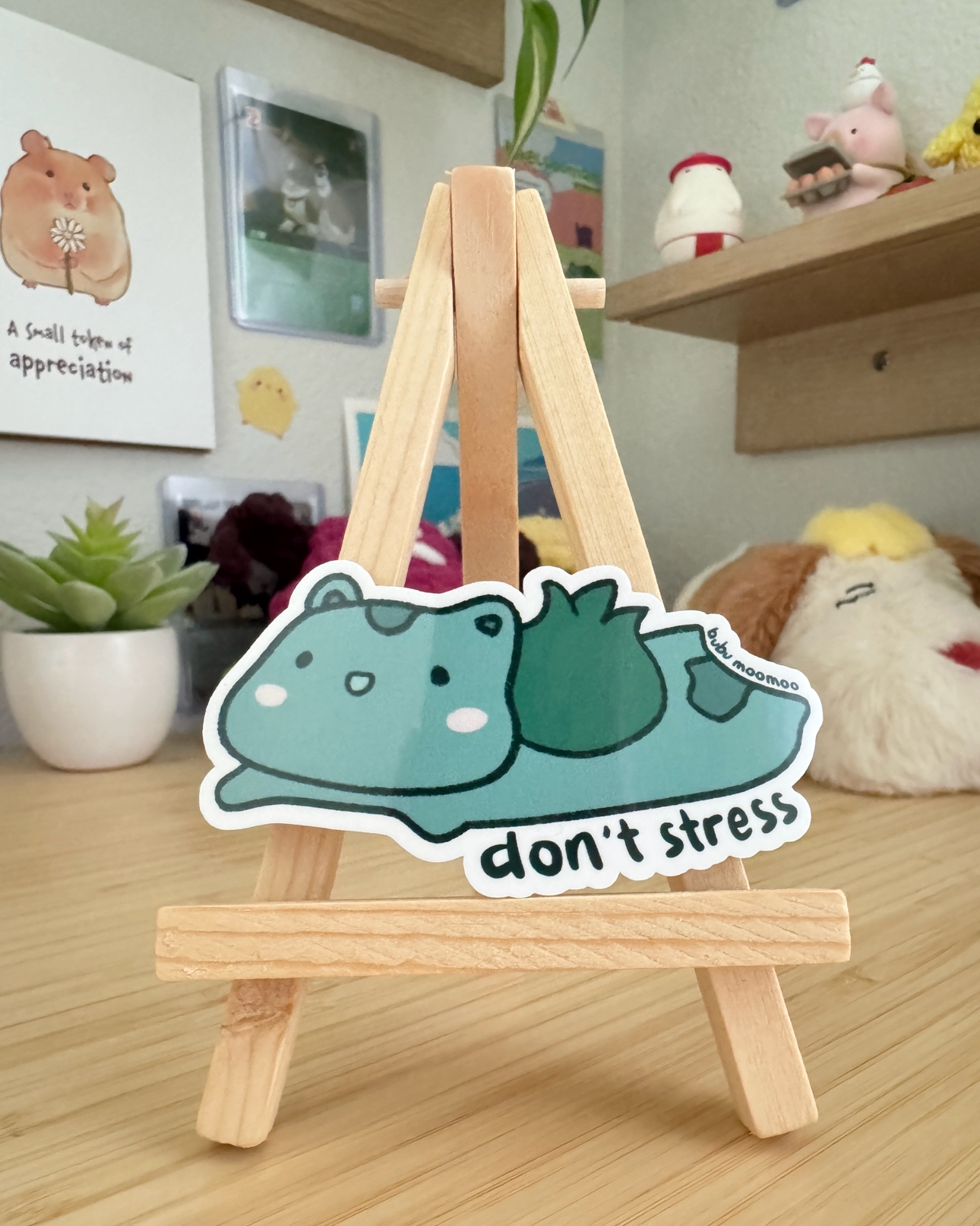 Small wooden easel with a cartoon sticker of bulbaboor with the words "don't stress" on a wooden desk. 