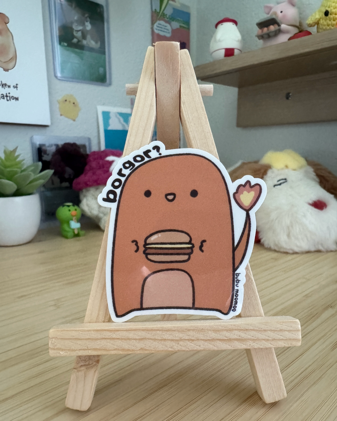 Small wooden easel with a cartoon character sticker of charburgur with the words "borgor?" on a wooden desk.
