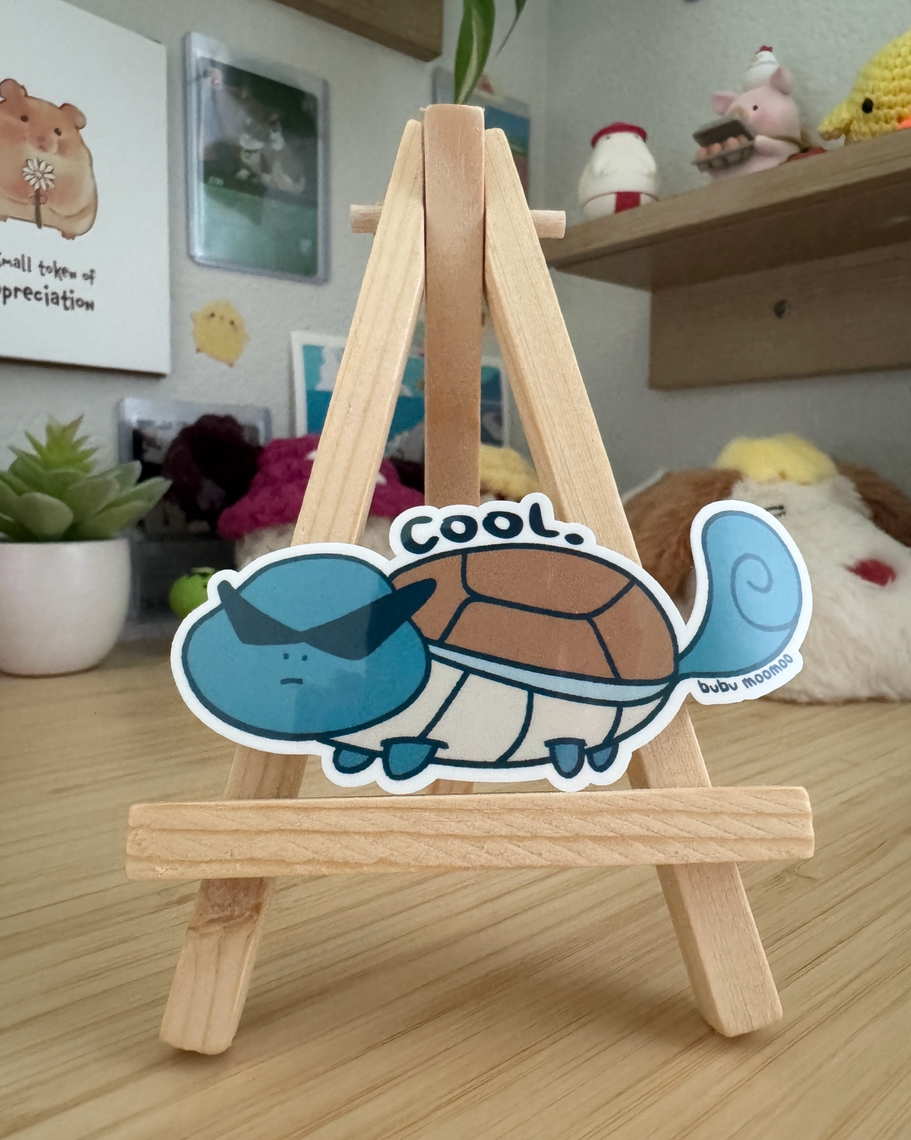 Small wooden easel with a cartoon character sticker of a turtle with the word "cool" on a wooden desk.